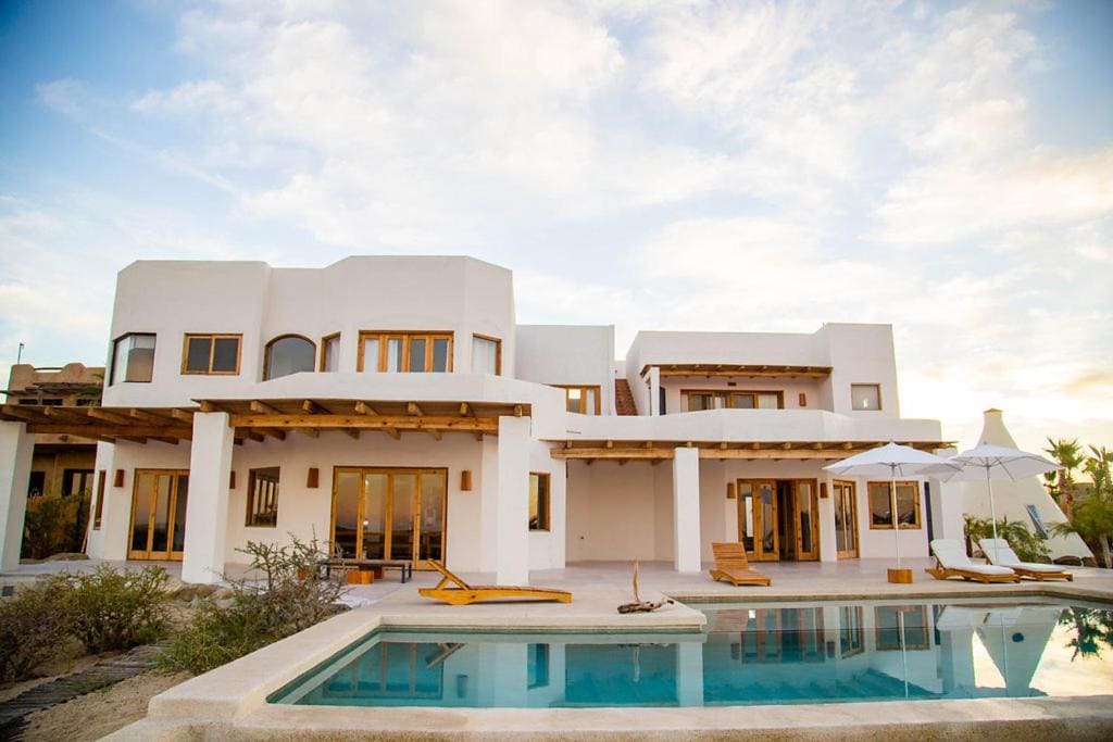 Where to Stay in Todos Santos, Mexico: Hotel & Area Guide 18 Elegant white stucco beachfront villa in Los Cabos, Baja California Sur, featuring wooden accents, a spacious patio with lounge chairs, umbrellas, and a private pool reflecting the soft glow of a sunset sky.