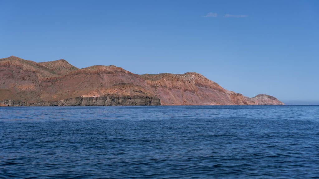 Our Day at Isla Espiritu Santo, La Paz: Sea Lions & Beaches 11 Rugged red and brown cliffs rise from deep blue water along the coastline of Isla Espiritu Santo in La Paz Baja California Sur under a clear sky.