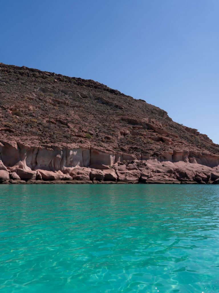 23 Best Things to Do in La Paz, Baja California Sur, Mexico 15 Bright turquoise water reflects sunlight in the foreground with rugged desert hills and layered rock formations along Isla Espiritu Santo in La Paz Baja California Sur.
