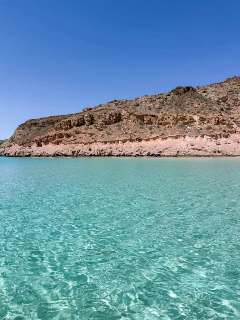 La Paz Mexico Travel Guide: Baja's Underrated Capital 20 Sunlit turquoise water ripples in the foreground with rocky desert cliffs and a cloudless blue sky at Isla Espiritu Santo in La Paz Baja California Sur.