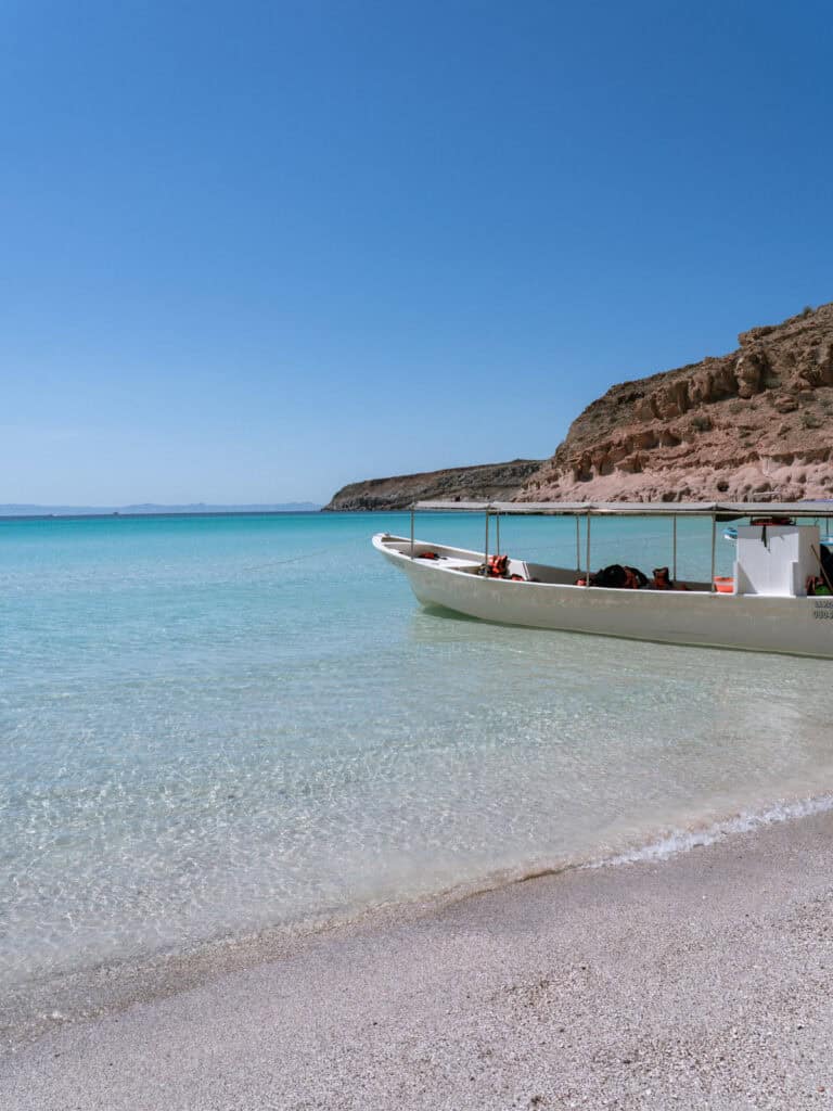 Our Day at Isla Espiritu Santo, La Paz: Sea Lions & Beaches 32 A long narrow boat is anchored in crystal clear shallow water beside a sandy beach with rugged hills behind it at Isla Espiritu Santo in La Paz Baja California Sur.