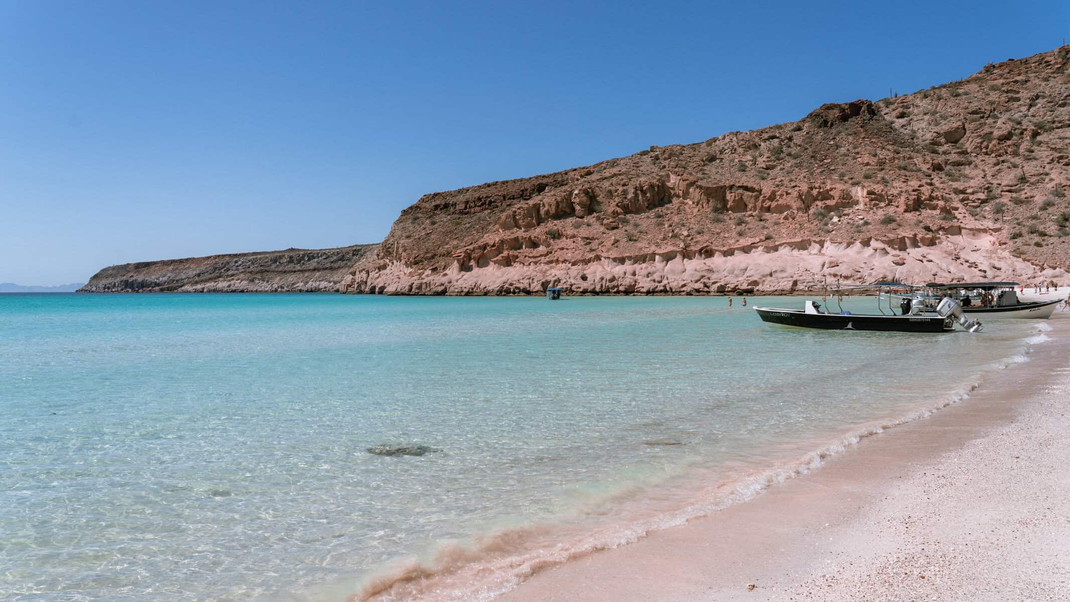 Our Day at Isla Espiritu Santo, La Paz: Sea Lions & Beaches 1 Several boats are anchored along a sandy beach with gentle waves and desert cliffs in the background at Isla Espiritu Santo in La Paz Baja California Sur.