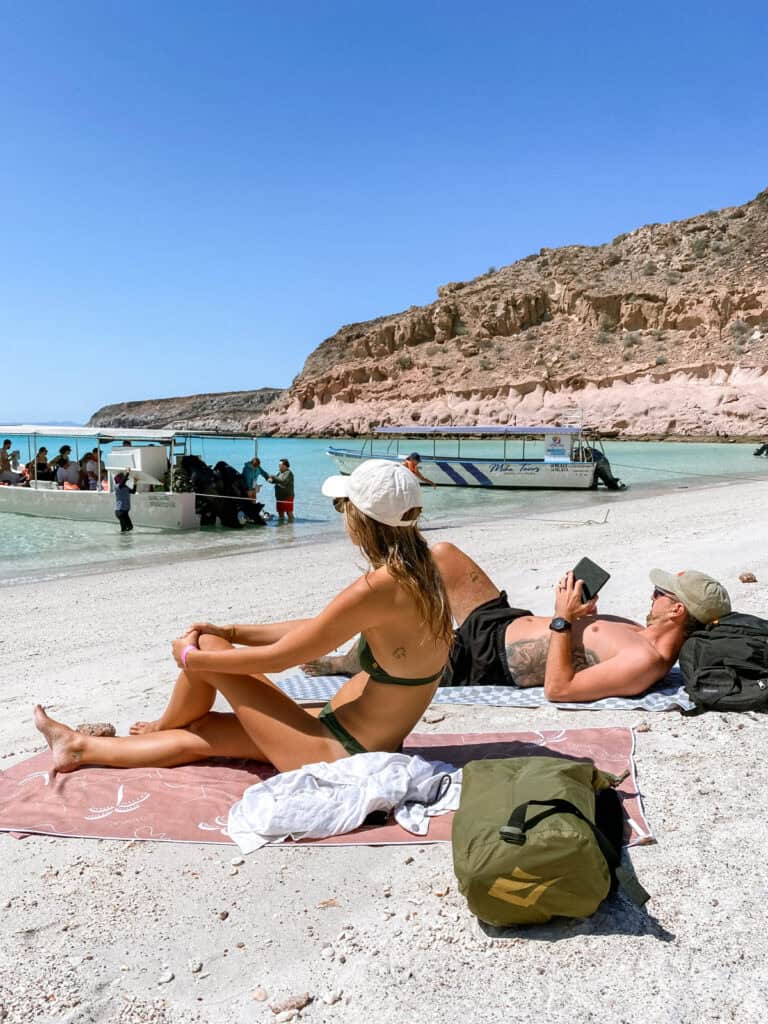 Our Day at Isla Espiritu Santo, La Paz: Sea Lions & Beaches 9 Sally sits on a beach towel facing the water while Brayden lies beside her looking at a phone as boats and people gather along the shoreline of Isla Espiritu Santo in La Paz Baja California Sur.