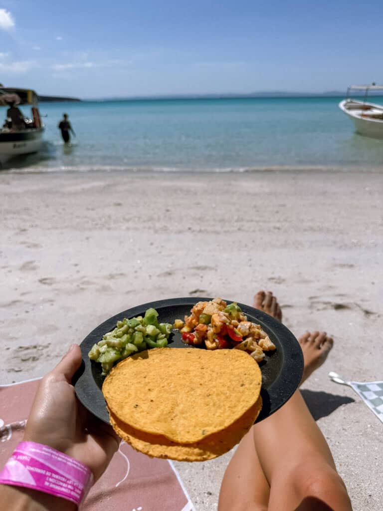 Our Day at Isla Espiritu Santo, La Paz: Sea Lions & Beaches 31 Sally relaxes on the beach holding a plate of tostadas topped with ceviche and avocado while facing calm turquoise water at Isla Espiritu Santo in La Paz Baja California Sur.