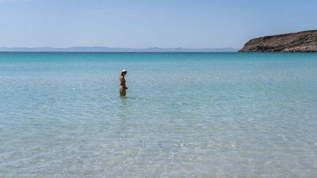 Our Day at Isla Espiritu Santo, La Paz: Sea Lions & Beaches 5 Sally stands waist deep in calm clear water looking toward the open sea under a bright blue sky at Isla Espiritu Santo in La Paz Baja California Sur.