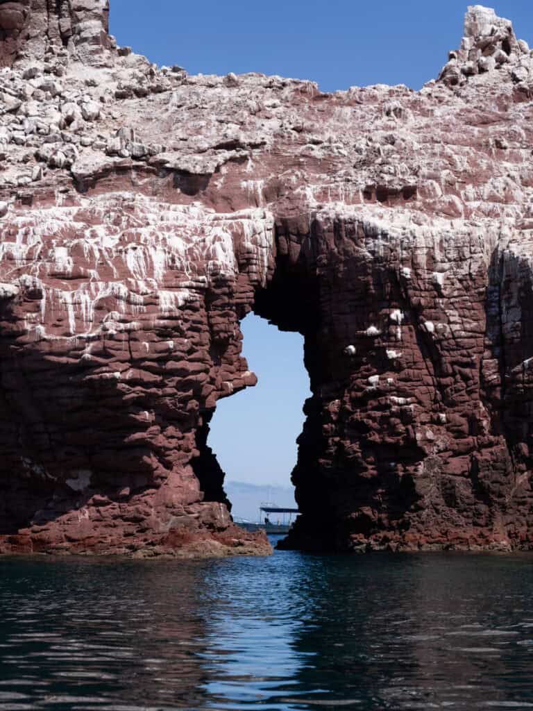 Our Day at Isla Espiritu Santo, La Paz: Sea Lions & Beaches 23 A dramatic natural rock arch rises from the water with layered red cliffs and a boat visible through the opening at Isla Espiritu Santo in La Paz Baja California Sur.