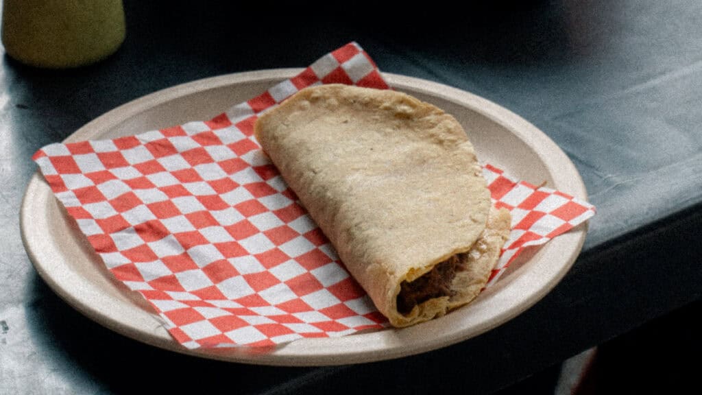 Mexico City Food Tour: Our Experience with Sherpa Food Tours 1 A half eaten stuffed pastry with flaky crust sits on a paper plate lined with red and white checkered paper on a metal counter during a Mexico City food tour.
