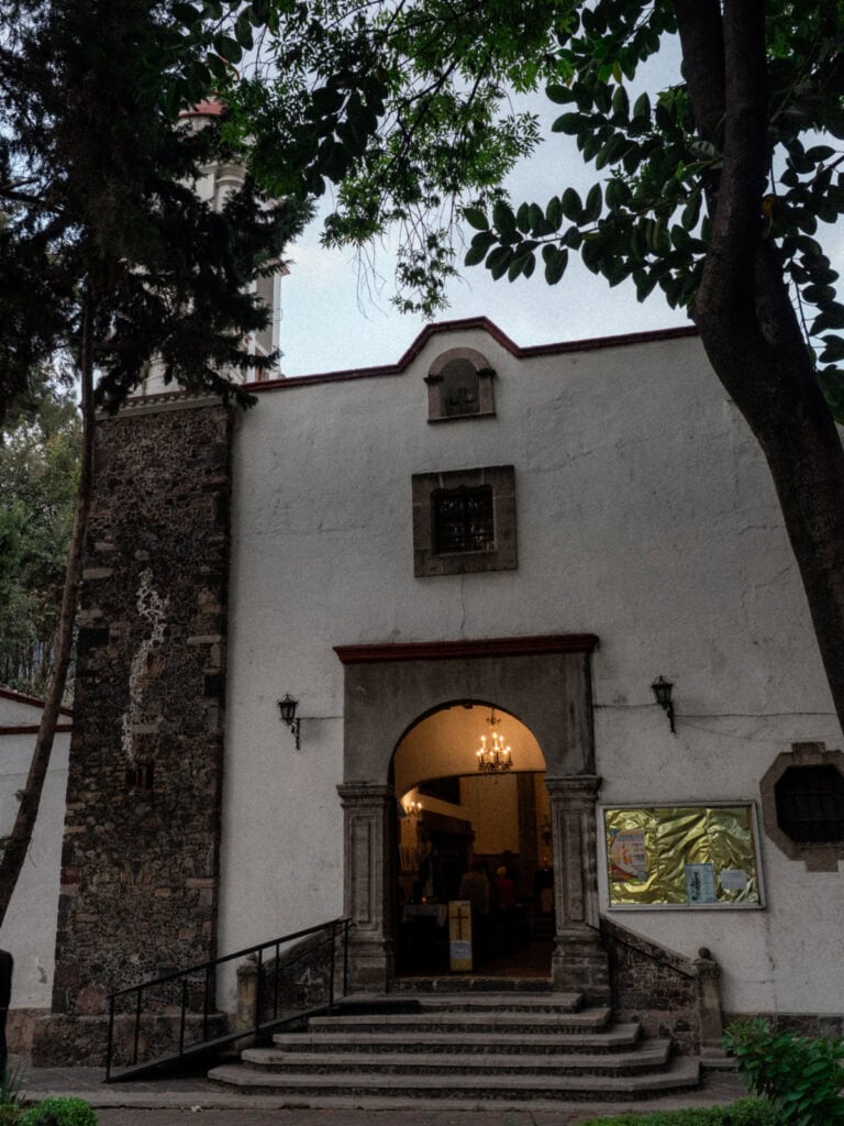 Mexico City Food Tour: Our Experience with Sherpa Food Tours 9 A small historic church in Mexico City is framed by leafy trees with a softly lit arched entrance and a stone bell tower rising behind it during a food tour stop.