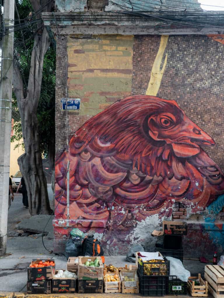 Mexico City Food Tour: Our Experience with Sherpa Food Tours 11 A large red bird mural painted on a weathered wall in Mexico City towers above crates of fresh produce like tomatoes and bananas with a street sign reading "Tizapán de Fonita" visible nearby.