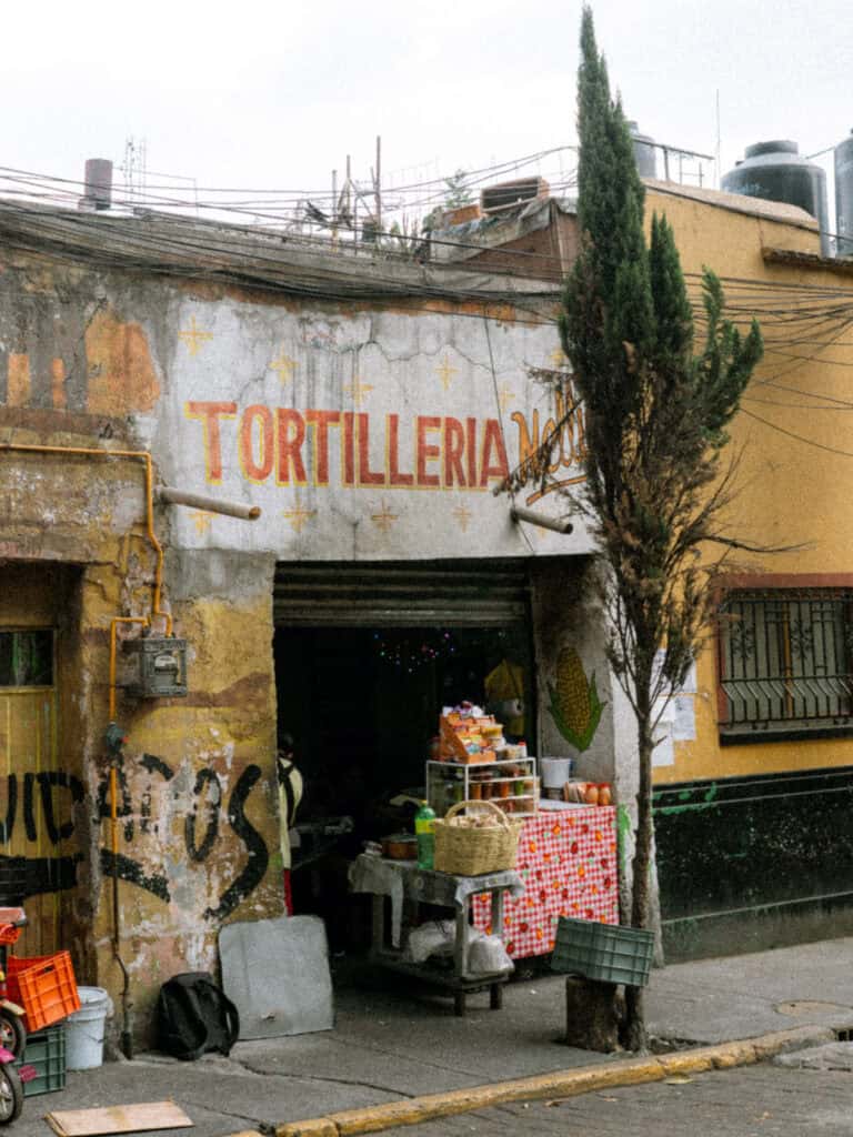 Mexico City Food Tour: Our Experience with Sherpa Food Tours 10 The exterior of a neighborhood shop in Mexico City shows a faded painted sign reading "TORTILLERIA" above the entrance with a small table of goods set outside along the street.