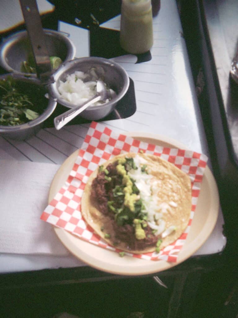 Mexico City Food Tour: Our Experience with Sherpa Food Tours 8 A taco topped with chopped onions, cilantro, guacamole, and sauce sits on a paper plate with red and white checkered paper beside small bowls of salsas and toppings during a Mexico City food tour.