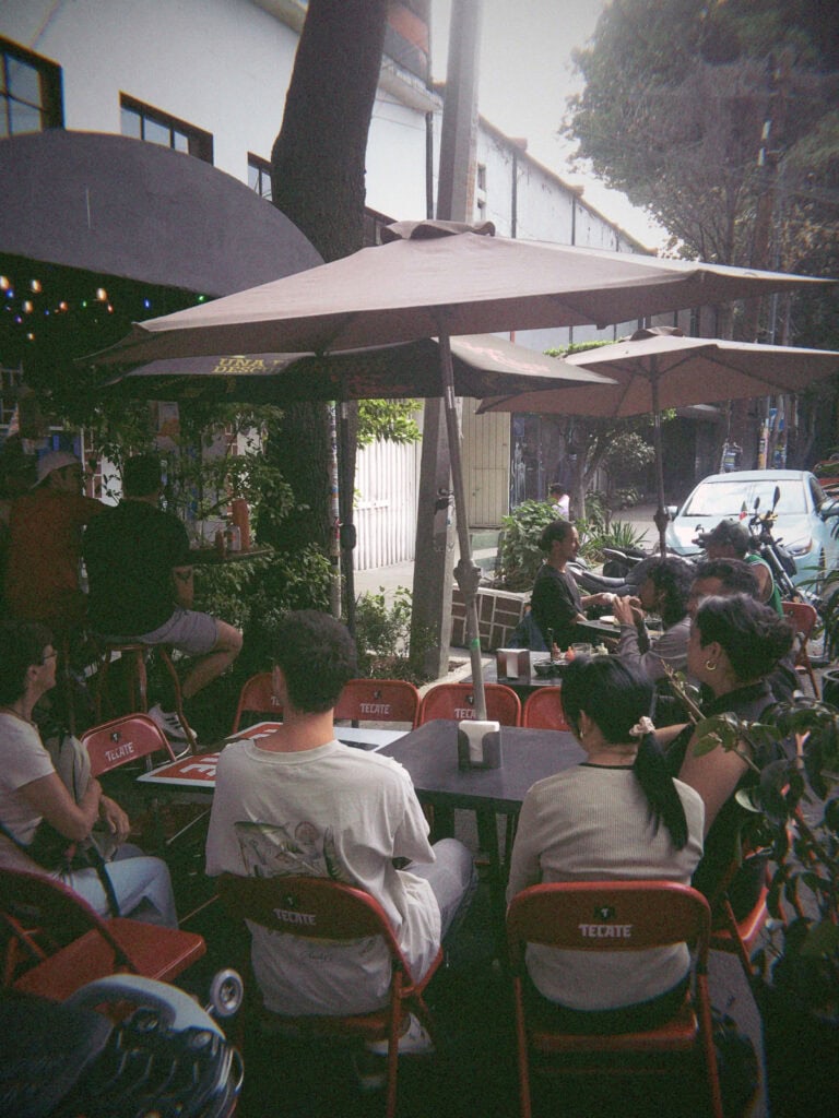 Mexico City Food Tour: Our Experience with Sherpa Food Tours 7 A group of people sit outdoors at a casual street food spot in Mexico City under large umbrellas on red chairs while listening to a guide during a food tour.