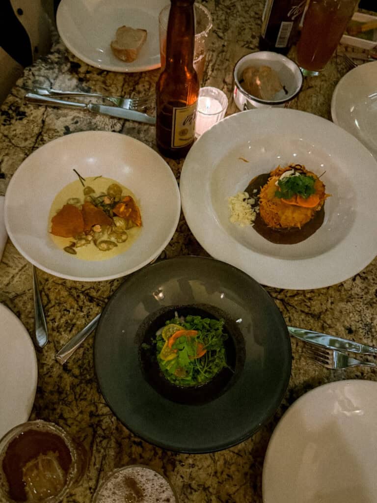Mexico City Food Tour: Our Experience with Sherpa Food Tours 19 Multiple plated dishes including a creamy dish with vegetables and a crispy topped entrée are arranged on a table with drinks and candles during a Mexico City food tour.