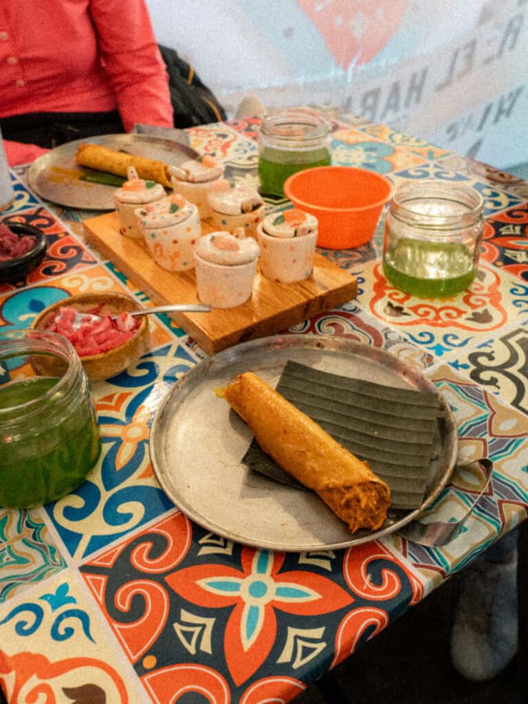 Mexico City Food Tour: Our Experience with Sherpa Food Tours 13 A close up of a rolled taco served on a plate with banana leaf alongside jars of green salsa, pickled onions, and small ceramic sauce containers on a colorful table in Mexico City.