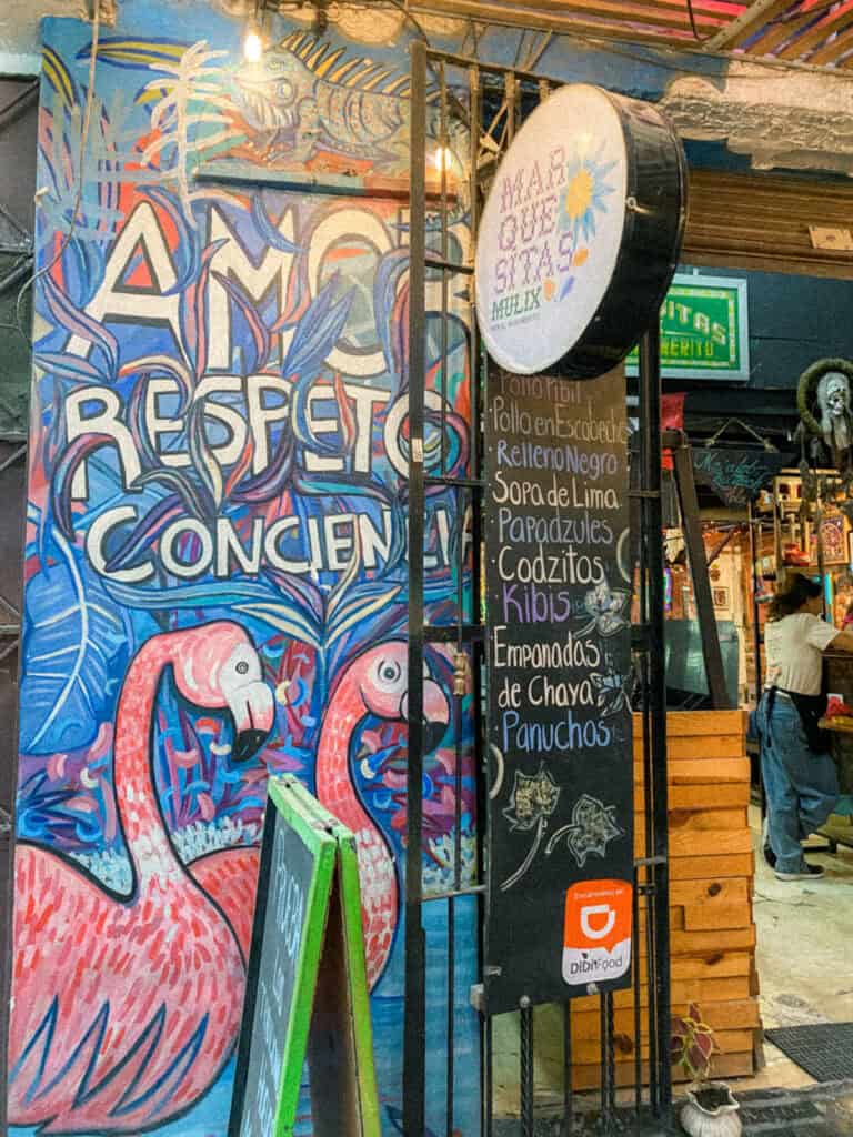 Mexico City Food Tour: Our Experience with Sherpa Food Tours 12 A bright mural in Mexico City featuring flamingos and bold text reading "AMOR RESPETO CONCIENCIA" stands beside a menu board listing dishes like "Sopa de Lima", "Papadzules", and "Empanadas de Chaya" during a food tour.