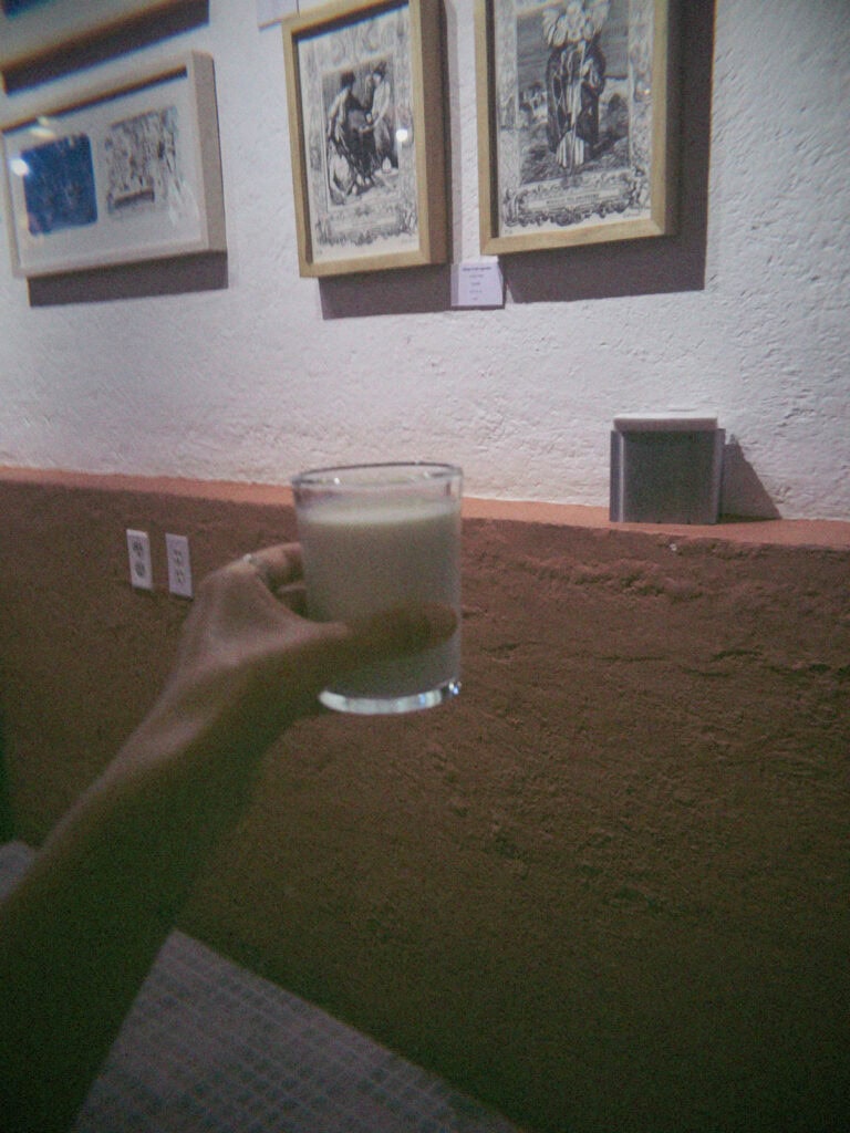Mexico City Food Tour: Our Experience with Sherpa Food Tours 15 A hand holds a glass of a creamy white drink in a dimly lit Mexico City restaurant with framed artwork hanging on the wall in the background.